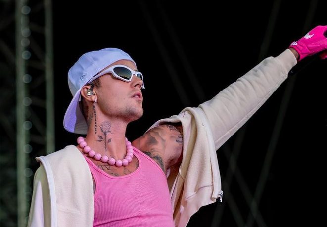 A young man with light skin tone stands on a stage with an outstretched left arm. He wears a backwards white ball cap, white sunglasses, a pink talk top and grey hoodie. Several tattoos can be seen on his arms and chest. He also wears a pink glove on his left hand. 