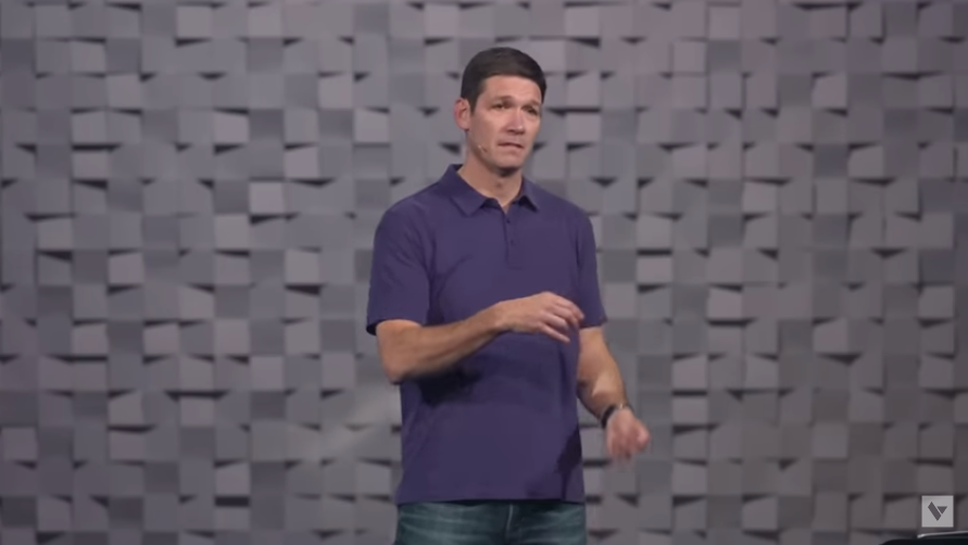 A man with light skin tone and short dark hair speaks from a stage and gestures with his right hand.