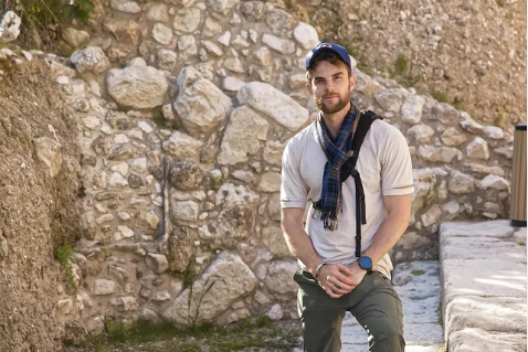 A man with lights skin tone stands on stone steps in front of an ancient stone wall. 
