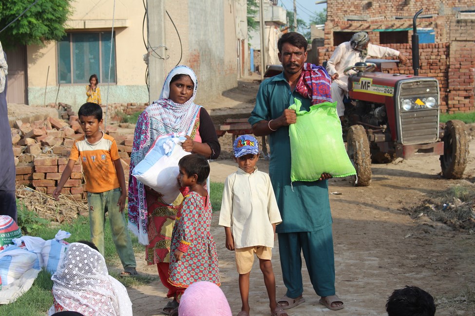 A family with dark skin tone stand with sacks of food. They stand on a dirt road with bricks stacked on the ground in the background. A man sits a small open cab red tractor in the background