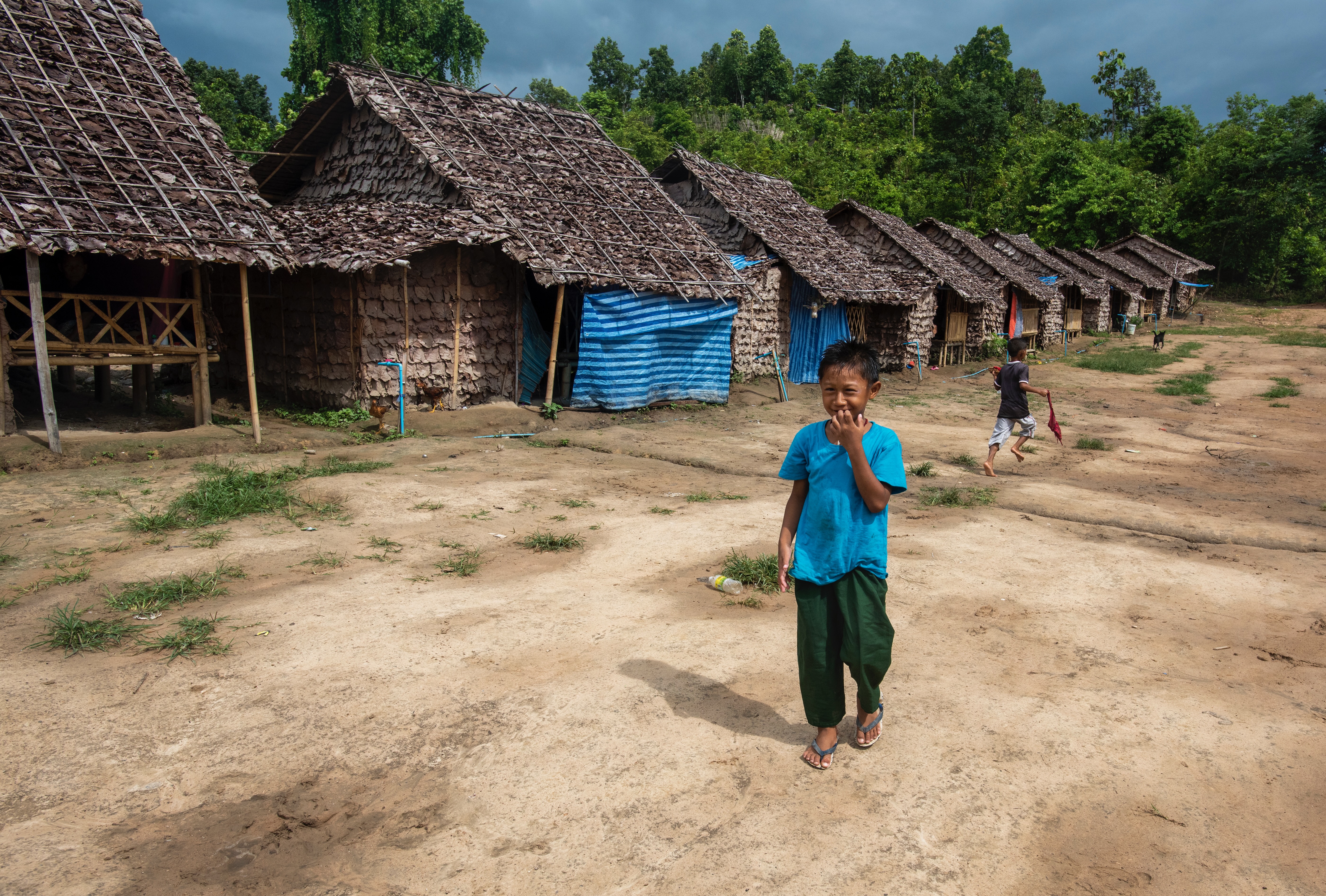 A young boy with medium skin tone and short dark hair walks on a dirt road in front of a row of huts with thatch roofs