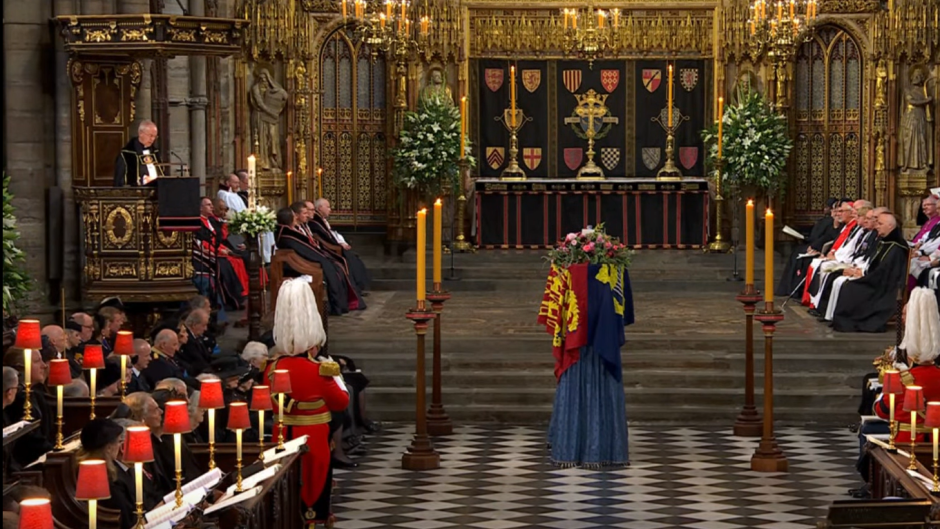 The sanctuary of a cathedral with a flag-draped coffin in front of the stairs up to the altar. A pries speaks from the side pulpit. 