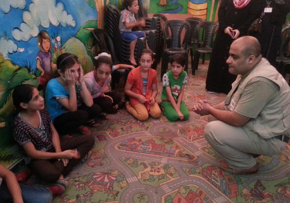 A mann squats down on the ground to talk with a group of children