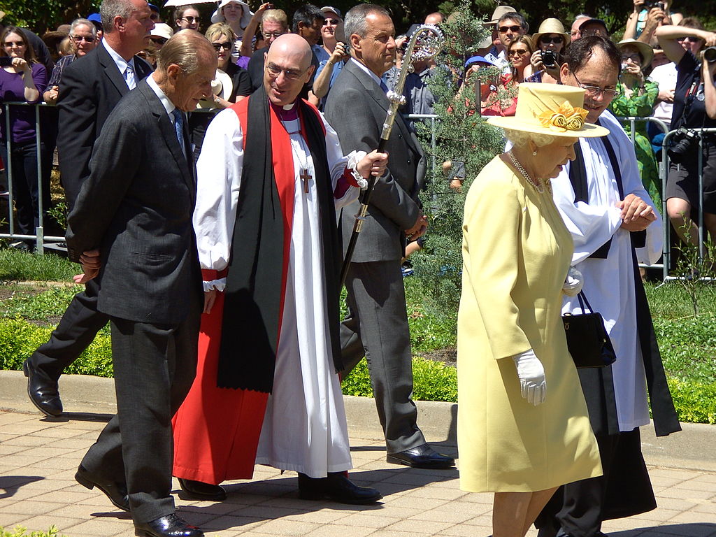 The queen wears a yellow dress and hat and walks outside with a priest. Her husband and another priest walk behind her