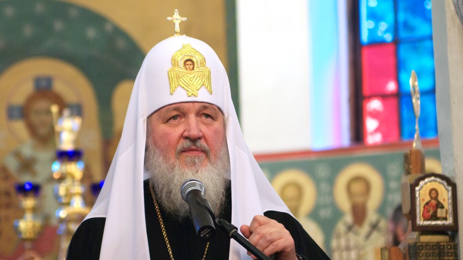 A man with a long white beard and a large white head cap stands in front of a microphone inside a church filled with iconography