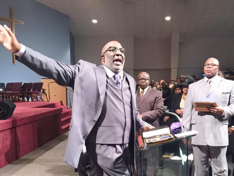 A man with dark skin tone holds his hand up and to his side as he gestures while preaching passionately from behind a pulpit. He wears a purple three-piece suit. Several congregants stand and listen in the background