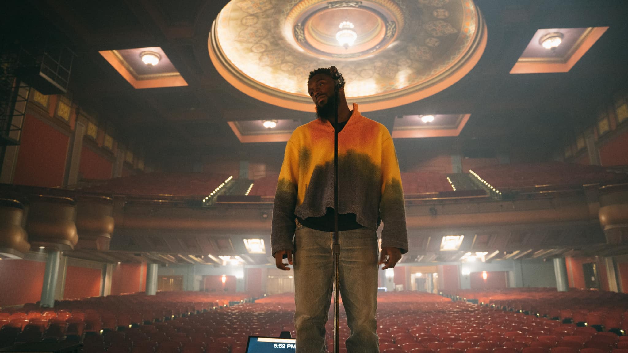 An upward shot of a man with dark skin tone standing on stage in an empty theatre.
