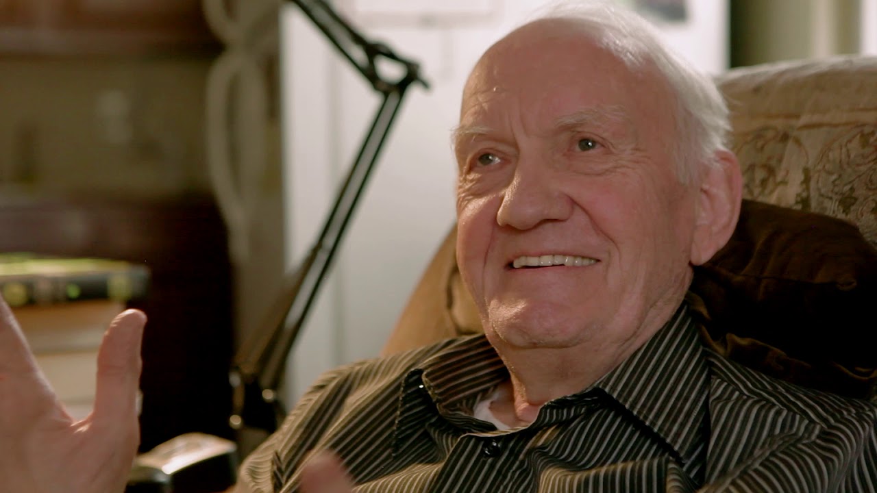 A balding man with grey hair smiles while sitting in a living room chair