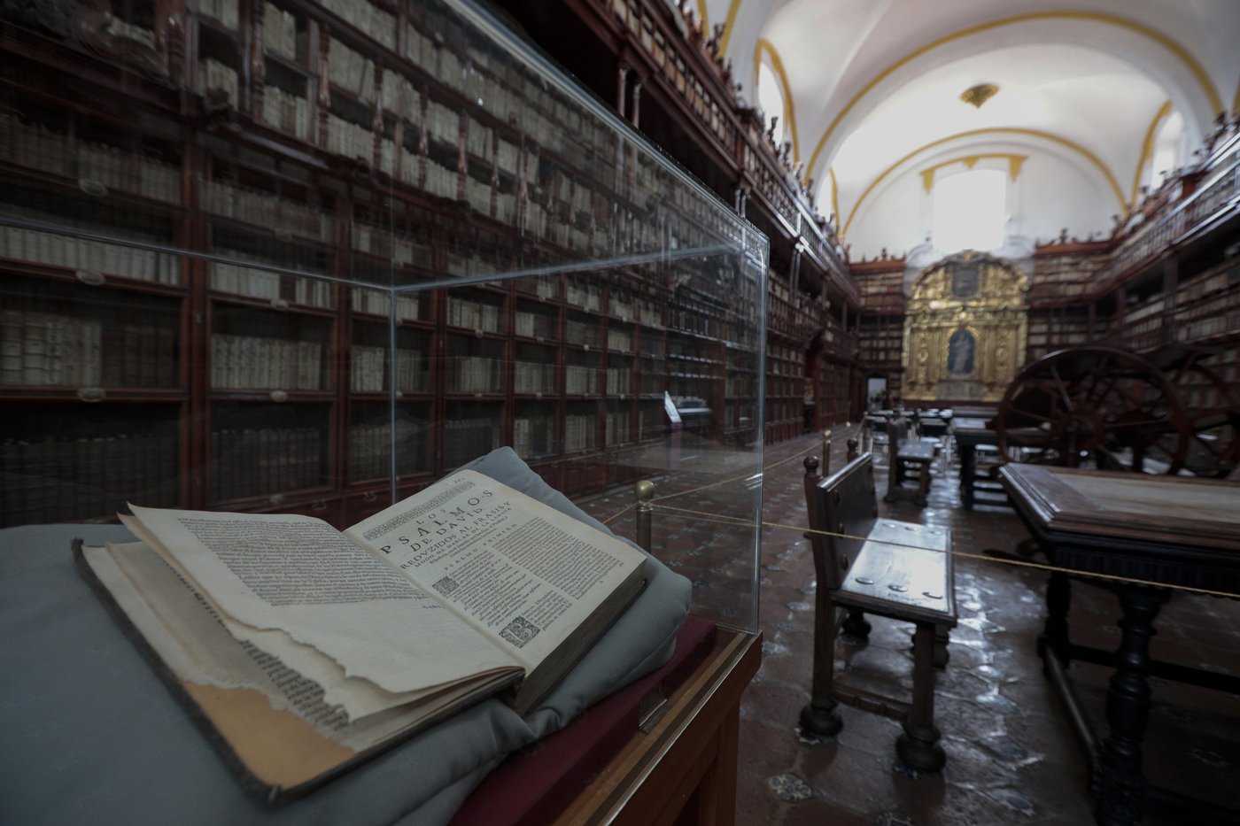 A opened book in a protective clear case is on display inside a large library with high wooden shelves filled with books