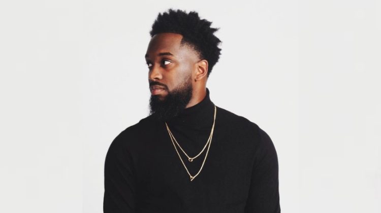 A man with dark skin tone and tall dark hair and dark beard wears a black turtle neck and 2 gold chains