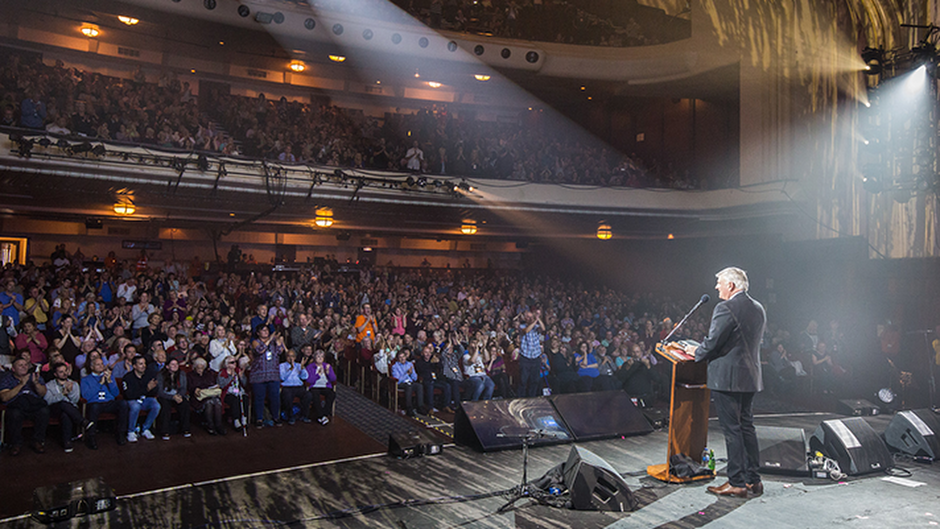 A shot from backstage of a man speaking behind a podium. A crowd of people look on in a theatre with a balcony