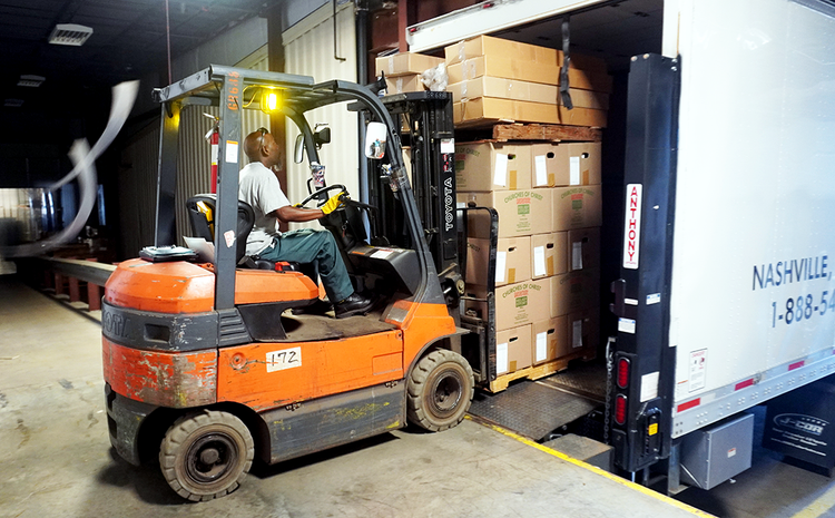 A man with dark skin tone drives an orange fork lift carrying a load of boxes on a pallet, backing it off a semi trailer and on to a loading dock