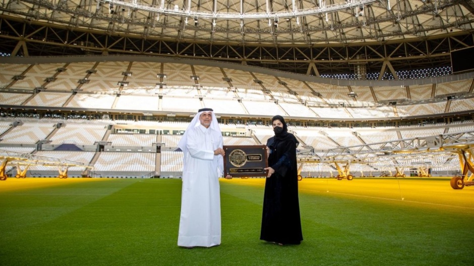 2 people stand each holding a side of a large plaque at centre field in a very large soccer stadium.