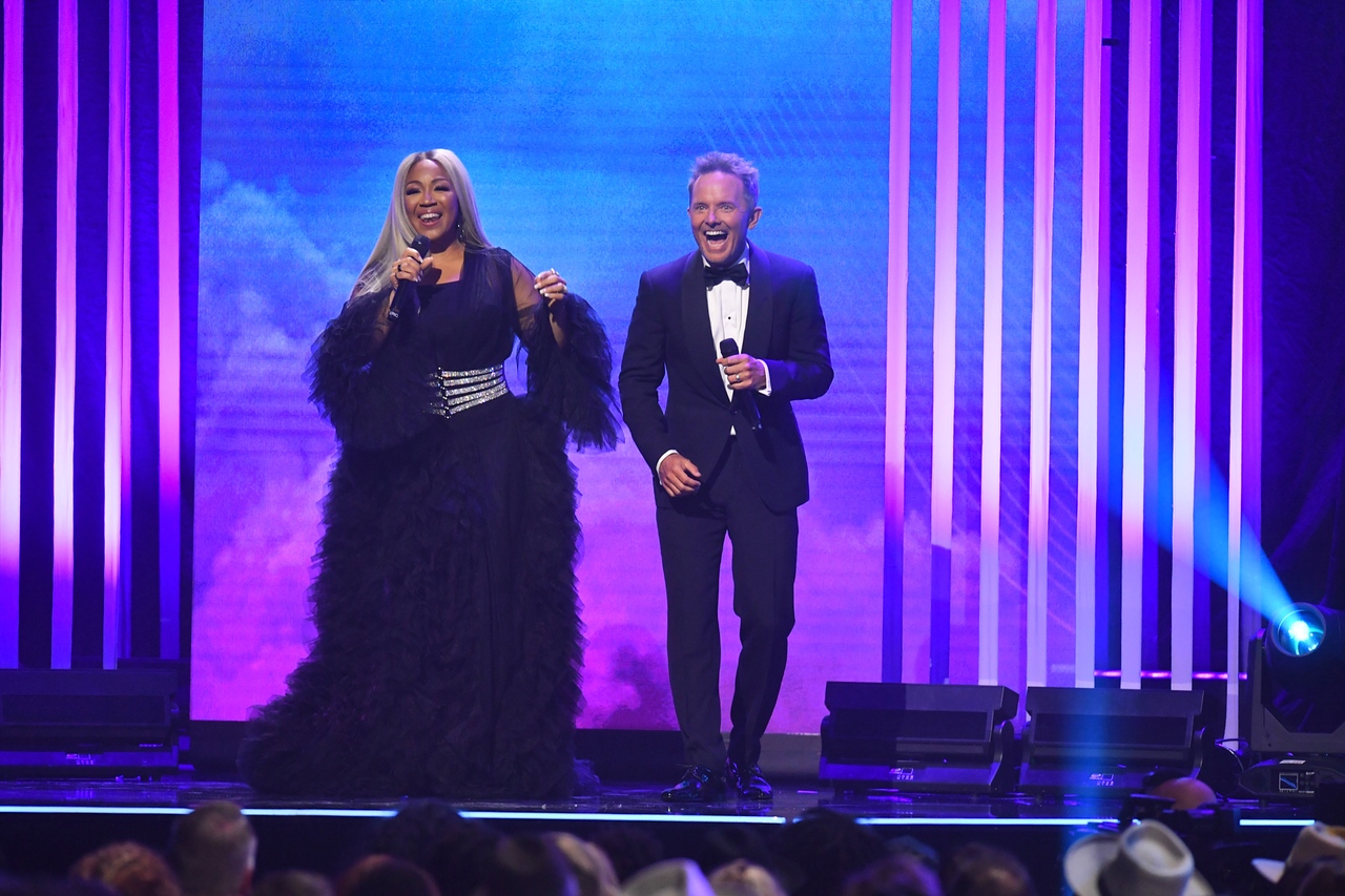 A woman with dark skin tone and long light hair wears a black dress. A man with light skin tone and short light hair wears a tuxedo. The stand on a stage in front of purple and blue lights and back dropss