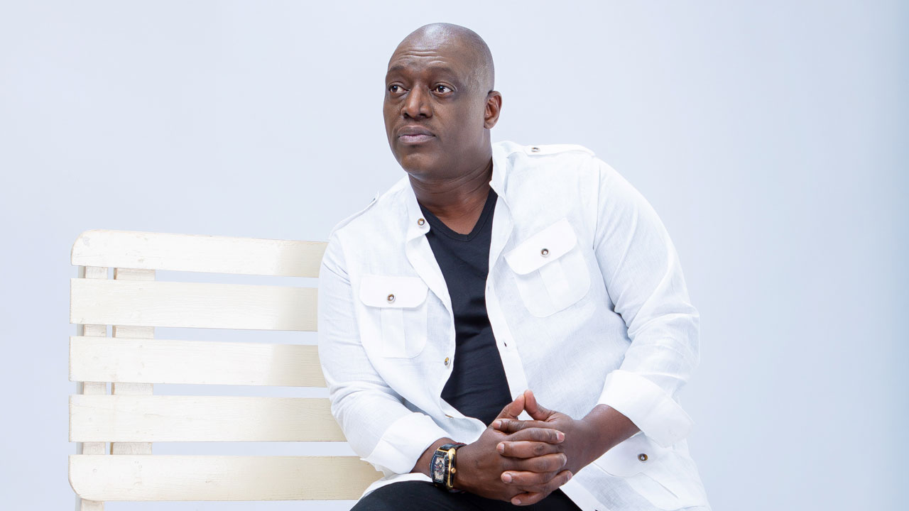 A publicity photo of a man with dark skin tone and a bald head. He sits on a white bench in front of a white backdrop. He wears a white jacket and black shirt.