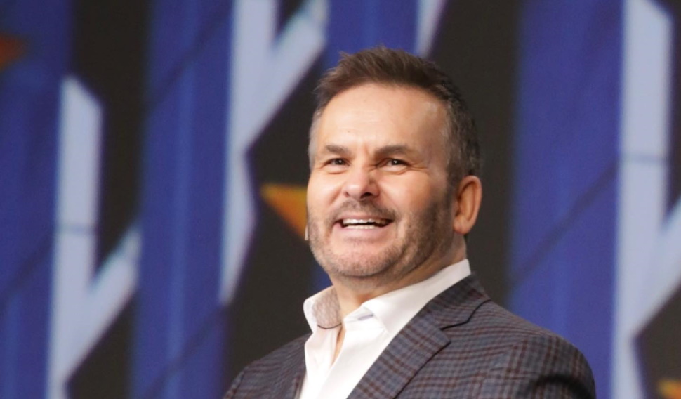 A man with light skin tone wears a dark sport coat and a white dress shirt and smiles as he speaks to a crowd