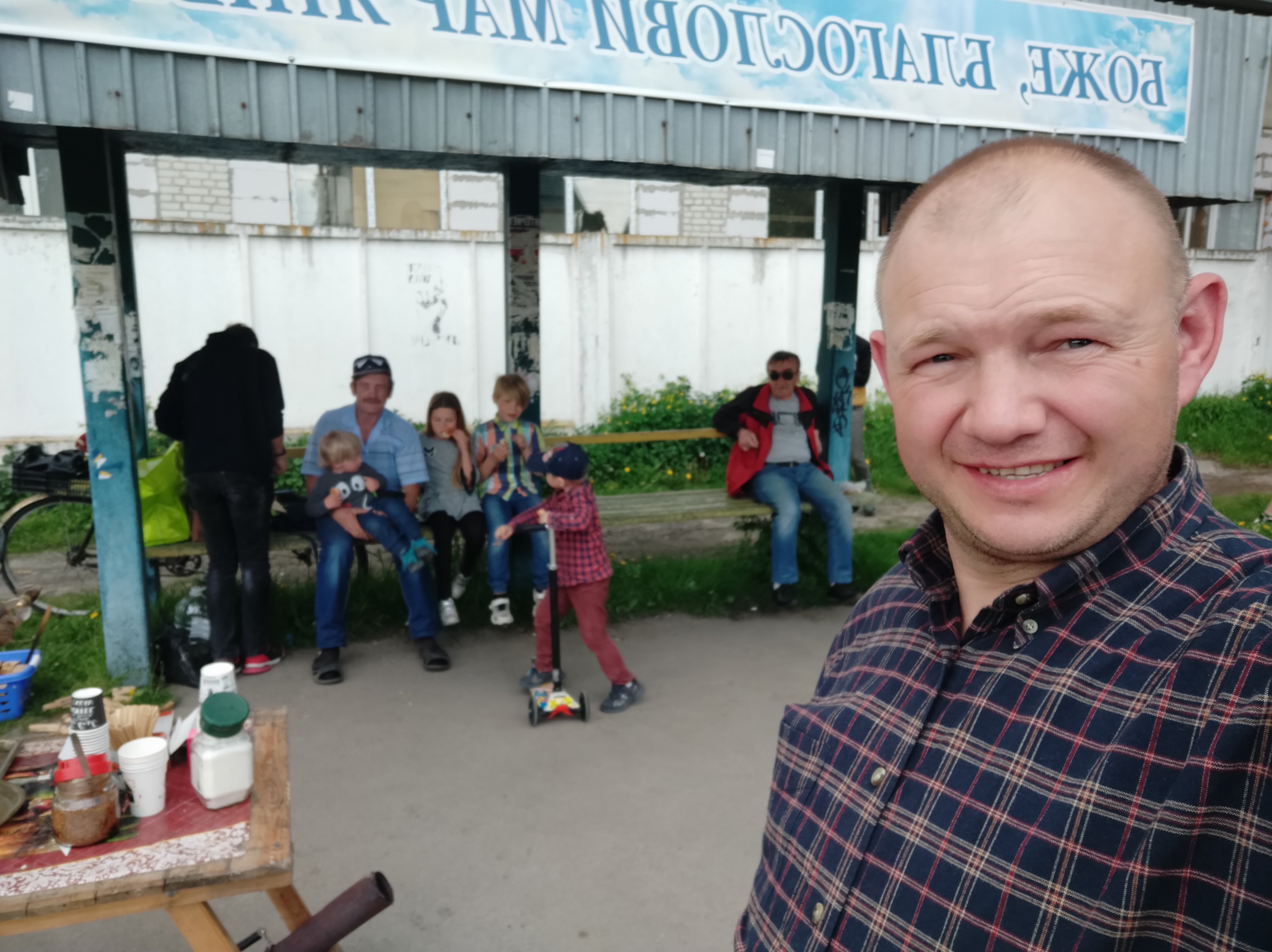 A man with light skin tone is in the foreground of a photo taking a selfie. A family made up of a man and 4 small children sit on a bench at a bus stop behind him; another man with light skin tone sits to their left