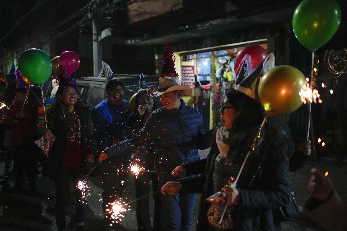 a group of young people hold sparklers and smile. One boy wears a cowboy hat and a girl wears a cone shaped party hat. They also hold colourful balloons