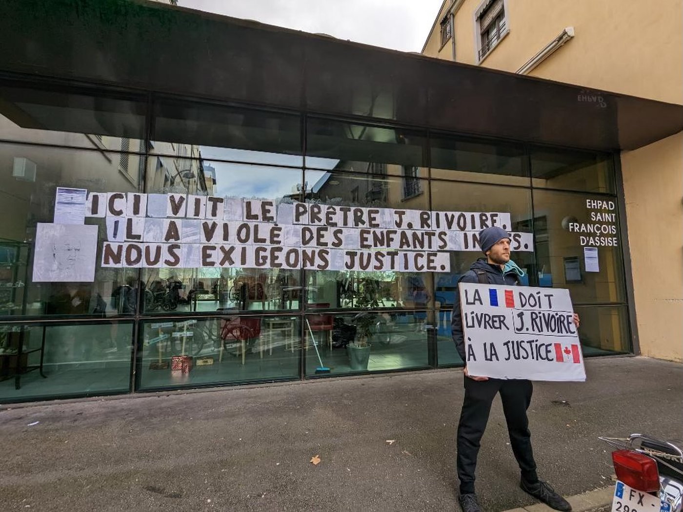 A person stands on a sidewalk on a sidewalk holding  a hand-written protest sign written in french which says "La [french flag] doit livrer J. Rivoire a la justice [canadian flag]"