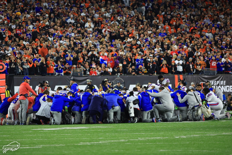 A large group of football players and staff kneel on the field in a circle and pray together.