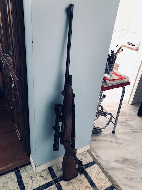 gun with scope used in shooting predators leaning against a wall as farmer prepares to head out.