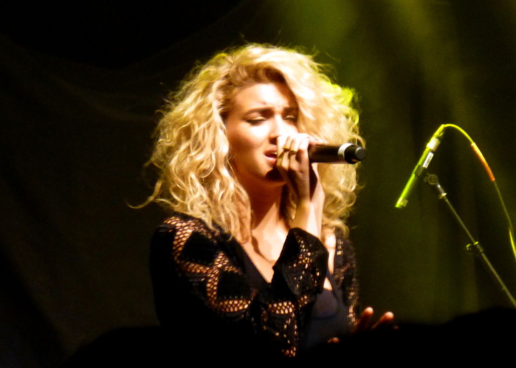 A woman with light skin tone and blonde hair sings into a hand held microphone
