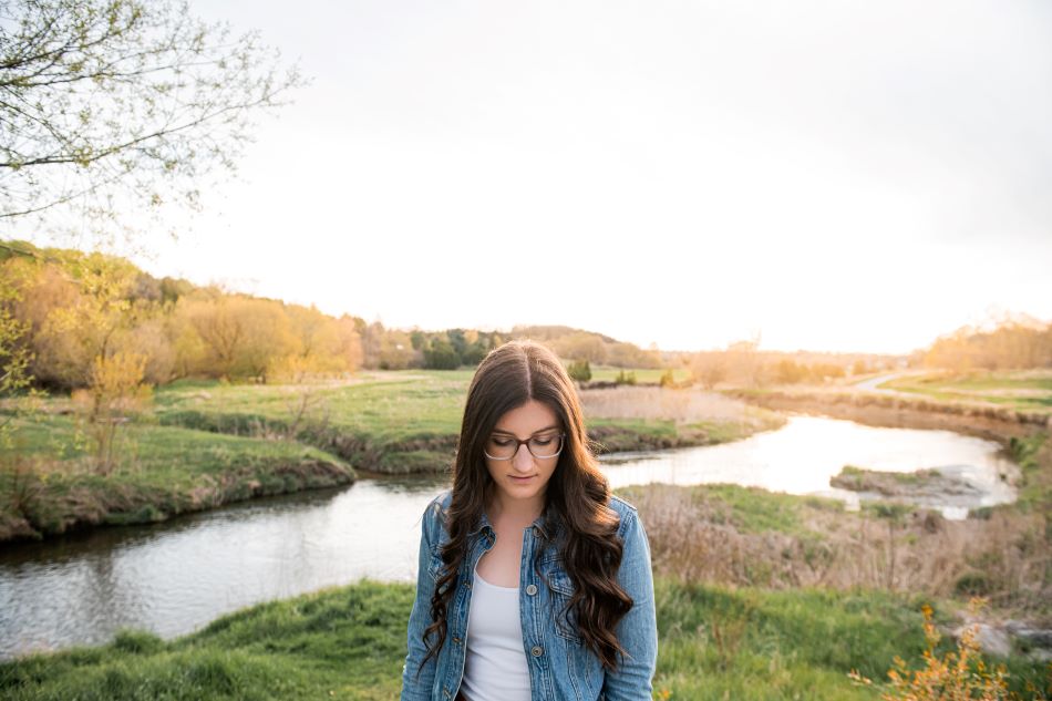 A young woman with light skin tone and long dark hair stands outside and gazes at the ground. A small river winds behind her through a wooded landscape