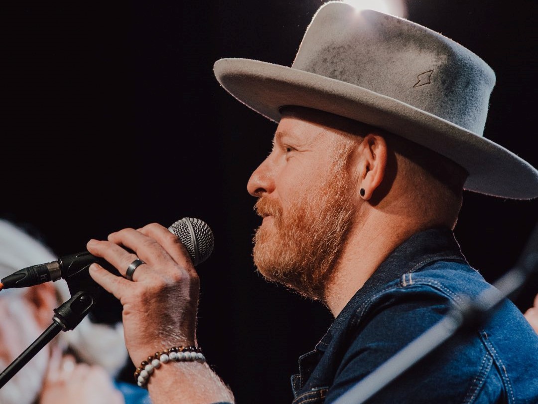A man with light skin tone and a red beard wears a wide brim hat and grabs a microphone with his hand