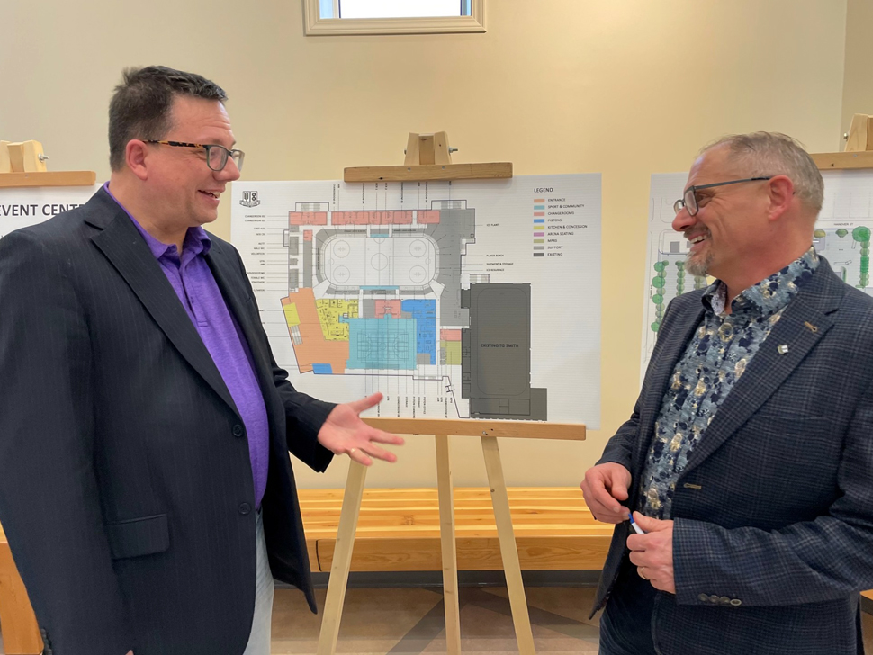 Two men in suit jackets standing in front of an architectural drawing of a large building. They are smiling, in conversation.