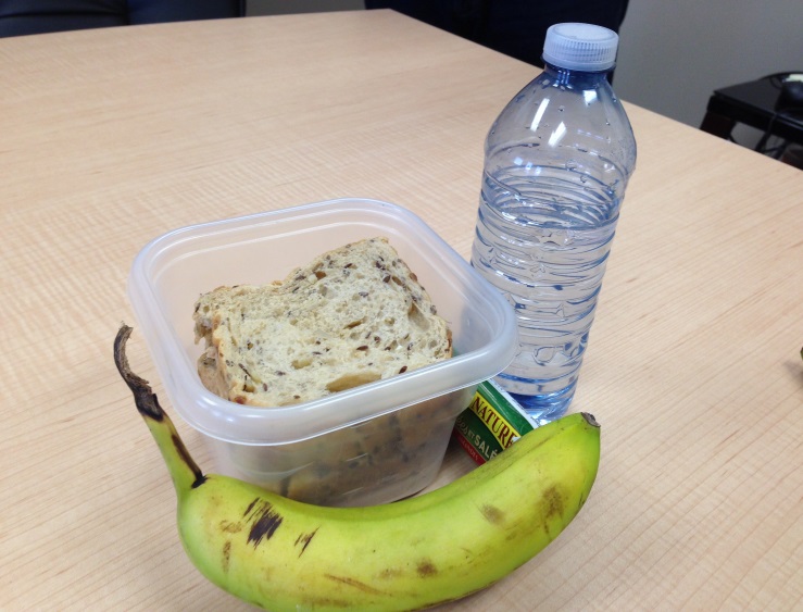 A sandwich, snack bar, banana and bottle of water sit atop a desk.