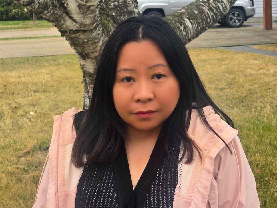 A woman with long black hair, wearing a black shirt and peach coloured jacket. In the background is a tree and grass.