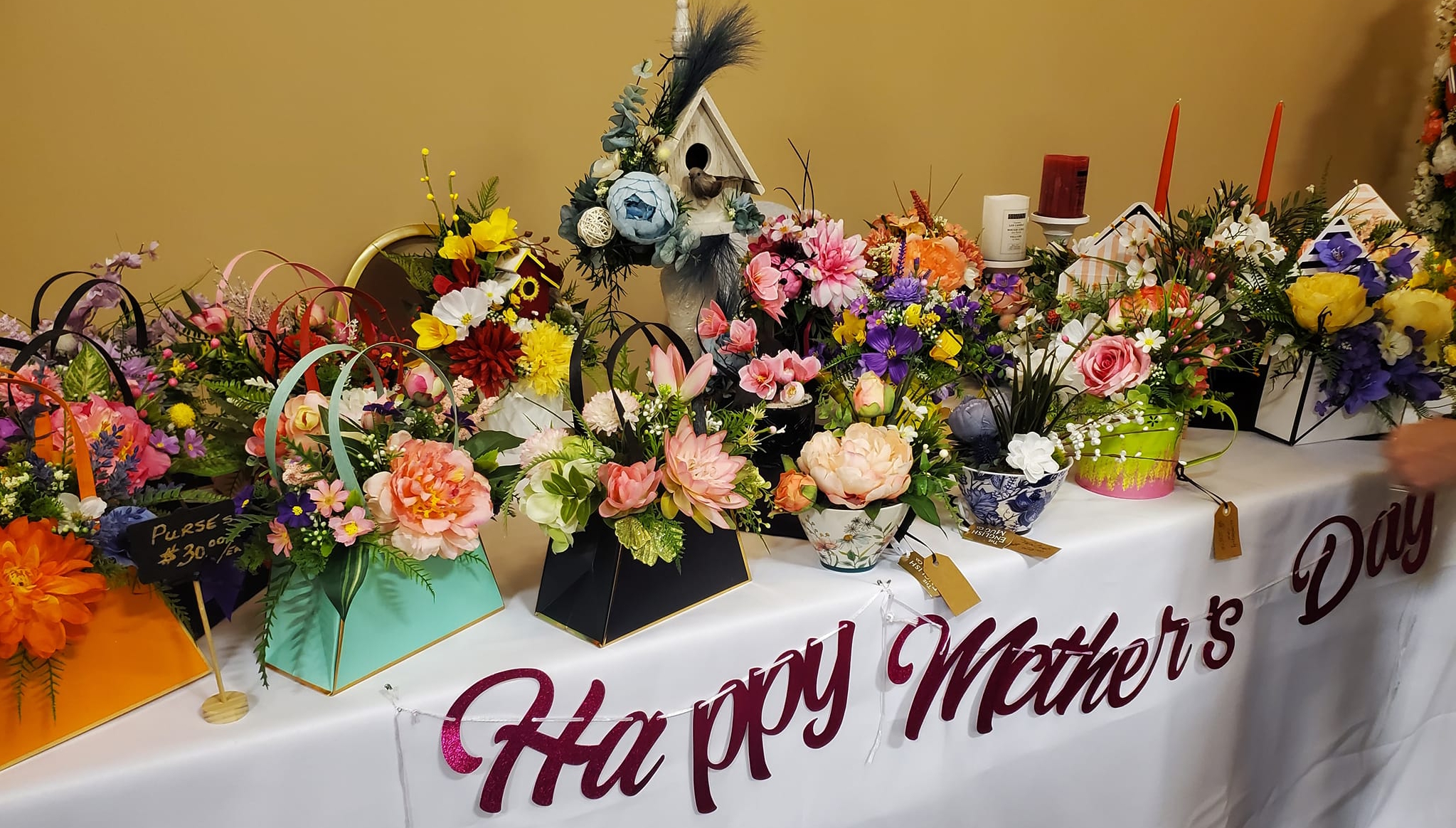 Colourful floral arrangements in baskets and teacups alongside other gifts on a vendor's table, featuring a 'Happy Mother's Day' banner, at the 2024 Portage Farmers' Market Mother's Day Market at Stride Place.