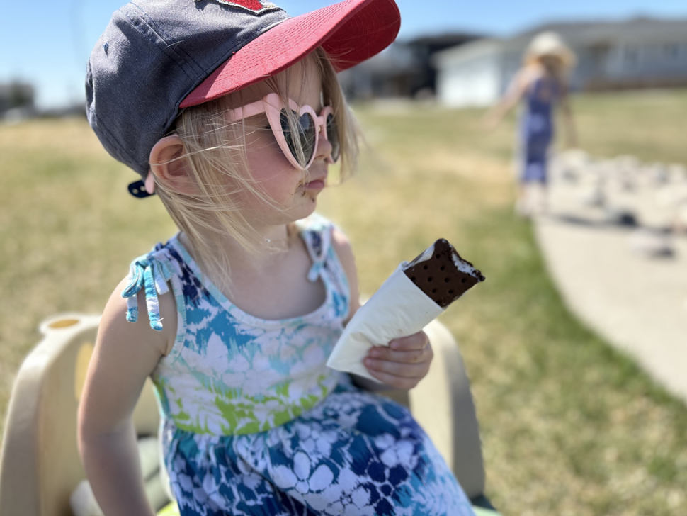 A female toddler with blond hair is wearing a ball cap and heart-shaped sunglasses. She has an ice cream sandwich in her hand. She sits on a chair on the grass. 