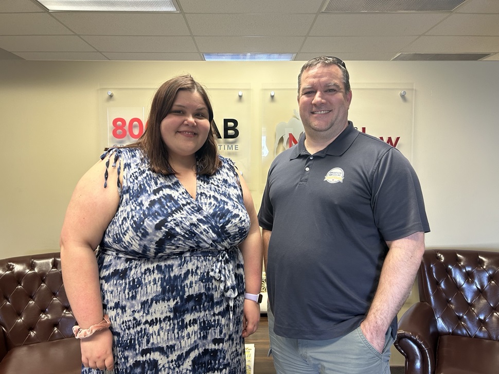 Two individuals stand side by side in an office setting. The person on the left wears a blue and white patterned dress, while the person on the right is dressed in a dark polo shirt and light-colored pants. The office features leather chairs and fluorescent lighting."