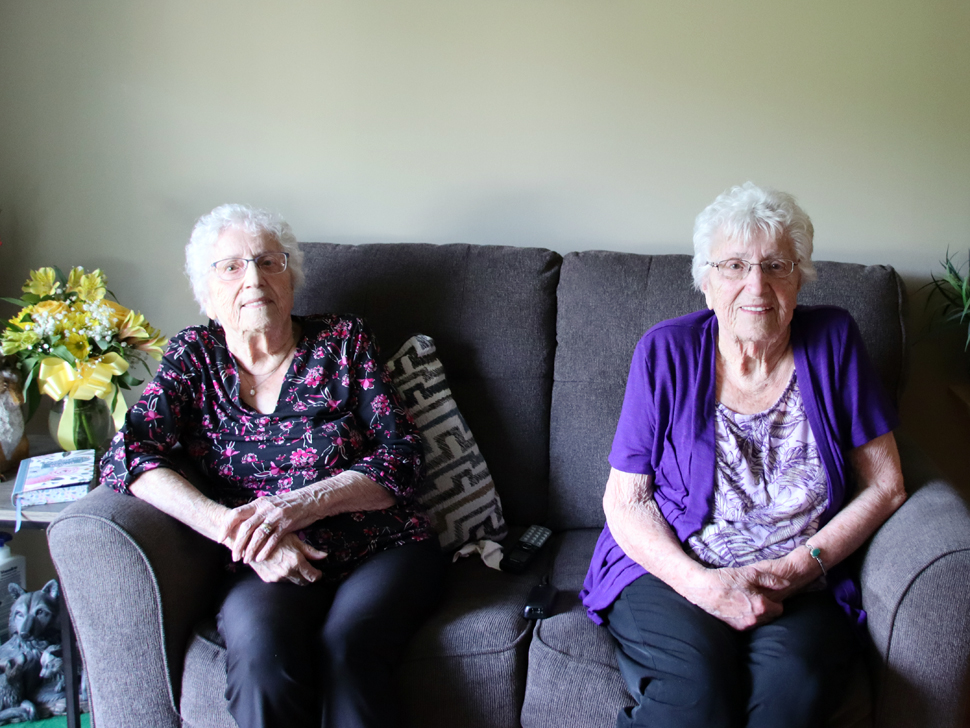 Two women sitting on a couch, they both have short grey hair, wear glasses, and are smiling.