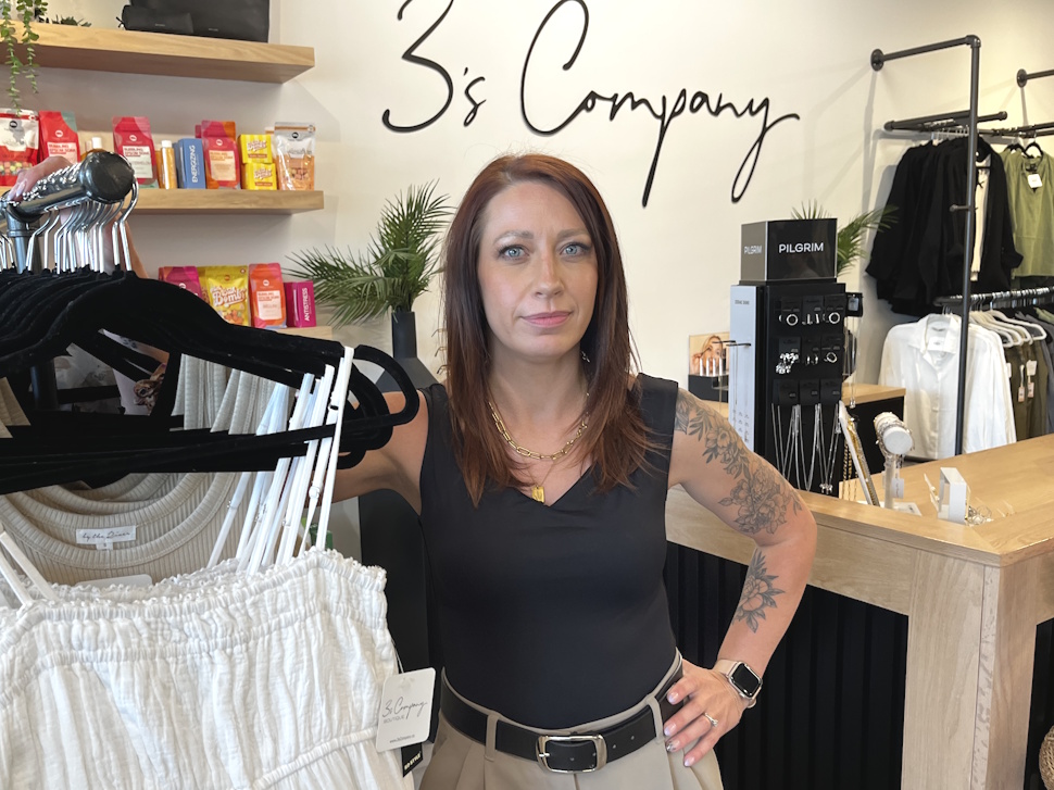 Janna Braaksma, owner of 3’s Company Boutique, stands inside her store looking directly at the camera, surrounded by clothing racks and boutique merchandise.