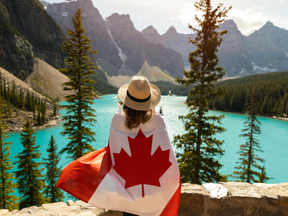 person standing on mountain top with Canadian flag wrapped around them. They are surrounded by trees, mountains and a bright blue lake