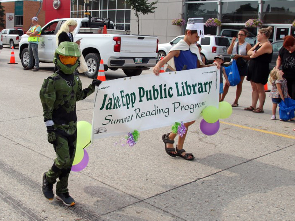 Two boys wearing costumes with headgear and capes, they are walking down the street holding a sign that reads Jake Epp Library.