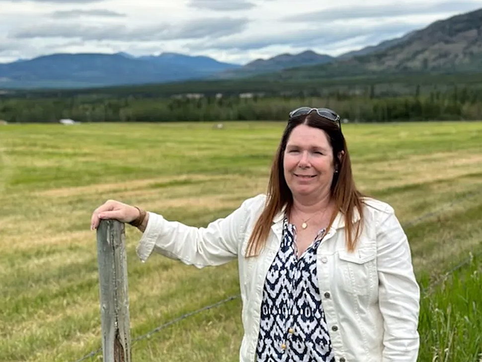 A woman with long brown hair standing by a fence post with a green field behind her.