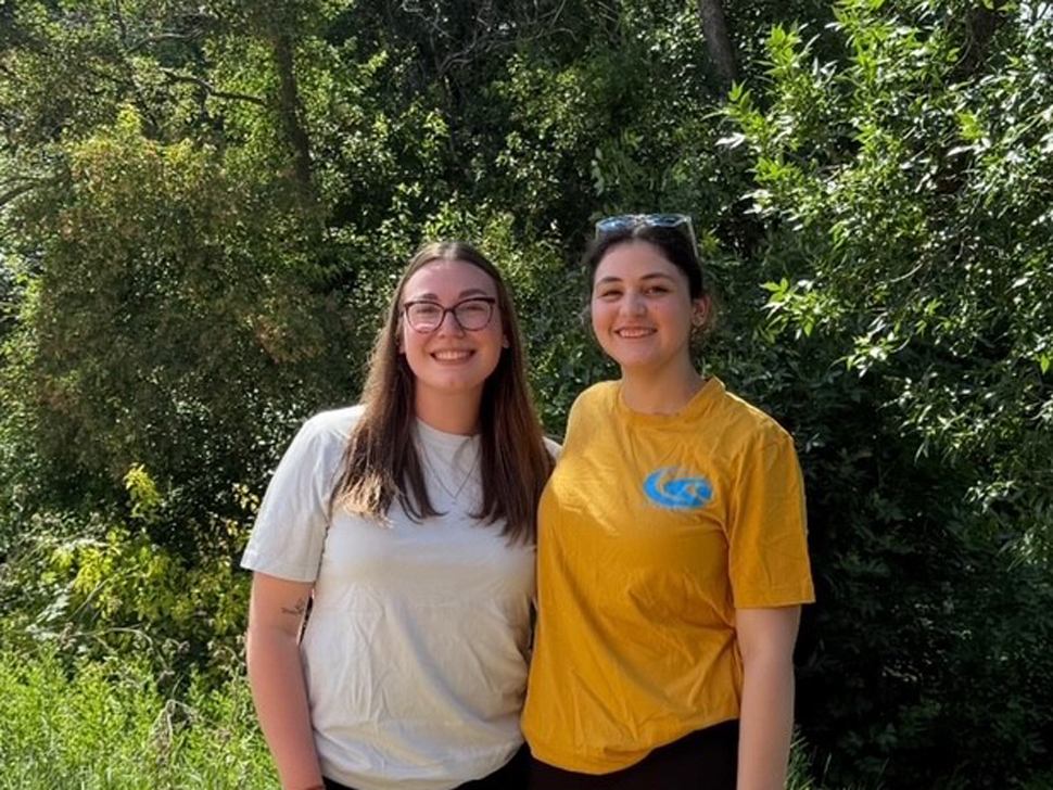 Two young ladies, both have long brown hair. There are standing in front of green bushes.