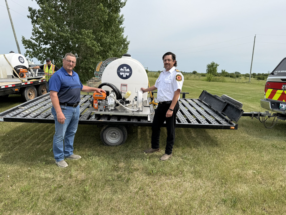Two men standing in front of a flatbed trailer that has a white circular tank on it. The grass is green.
