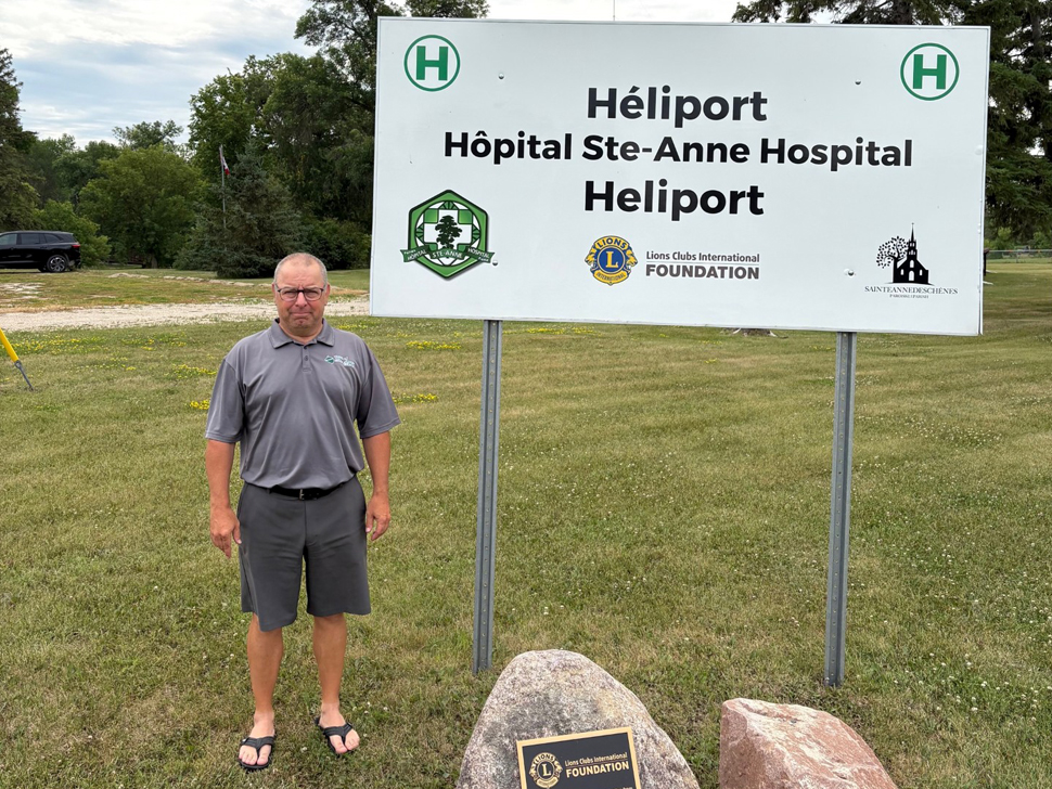 A man wearing a grey golf shirt and grey walking shorts standing beside a sign for the Ste. Anne Hospital Heliport.