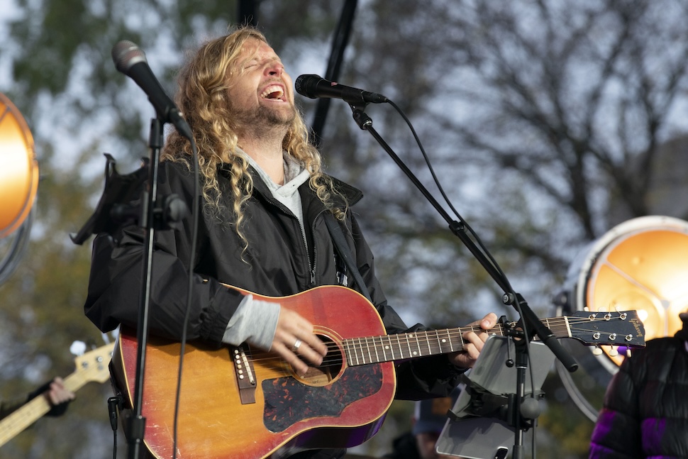 A man with long hair plays a guitar and sings into a microphone at an outdoor event