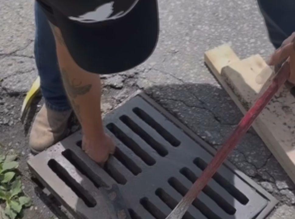 A man bends down and pulls up on a metal grate in the roadway with one hand while prying with a metal bar in the other hand