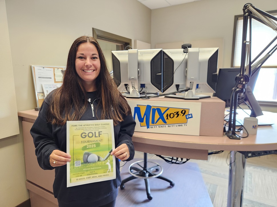 A person with long hair holds up a flyer promoting the 2025 Fore the Athletes Golf Classic.  Behind them, a radio studio is visible with computer monitors and a MIX 103.9 FM sign.