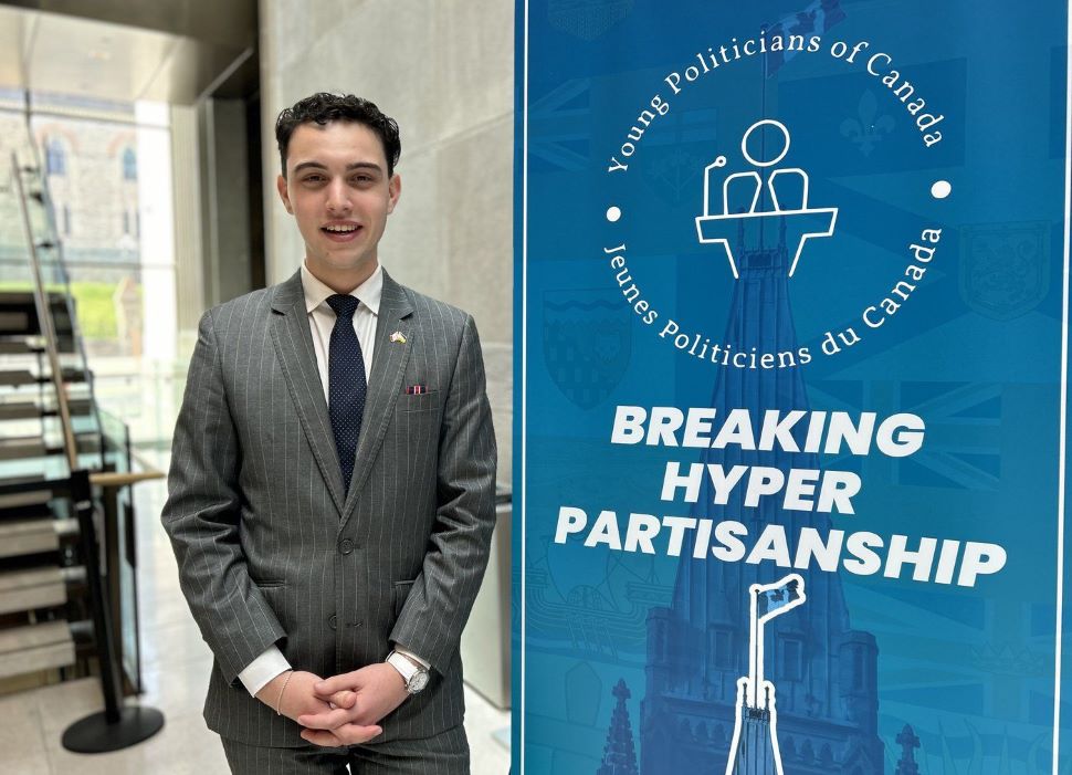 A young man stands next to a blue banner that shows a young politicians of Canada logo and says "Breaking Hyper Partisanship"
