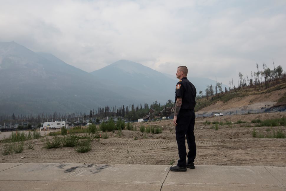 A man in a black firefighter's dress uniform stands on a sidewalk with mountains in the background