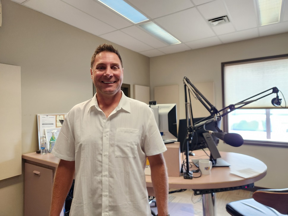 A person in a white short-sleeved shirt stands inside a radio broadcast studio.