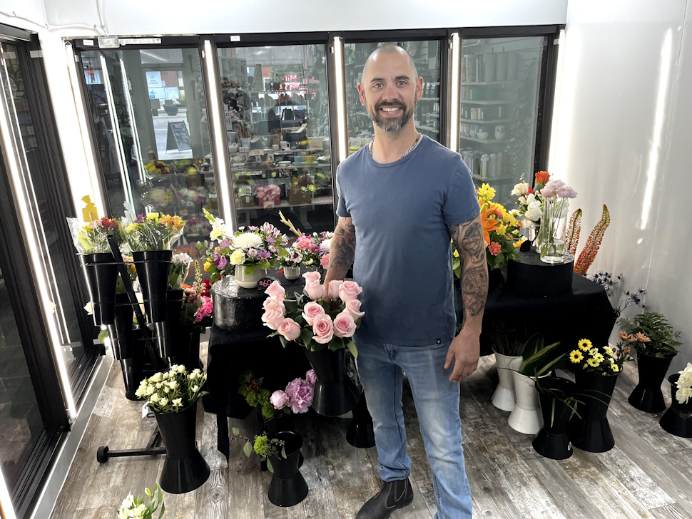 Kyle Pettinger inside the newly renovated walk-in cooler at Mil-Jeanne Flowers & Axcenz, holding a bouquet of fresh flowers with shelves of vibrant blooms in the background.