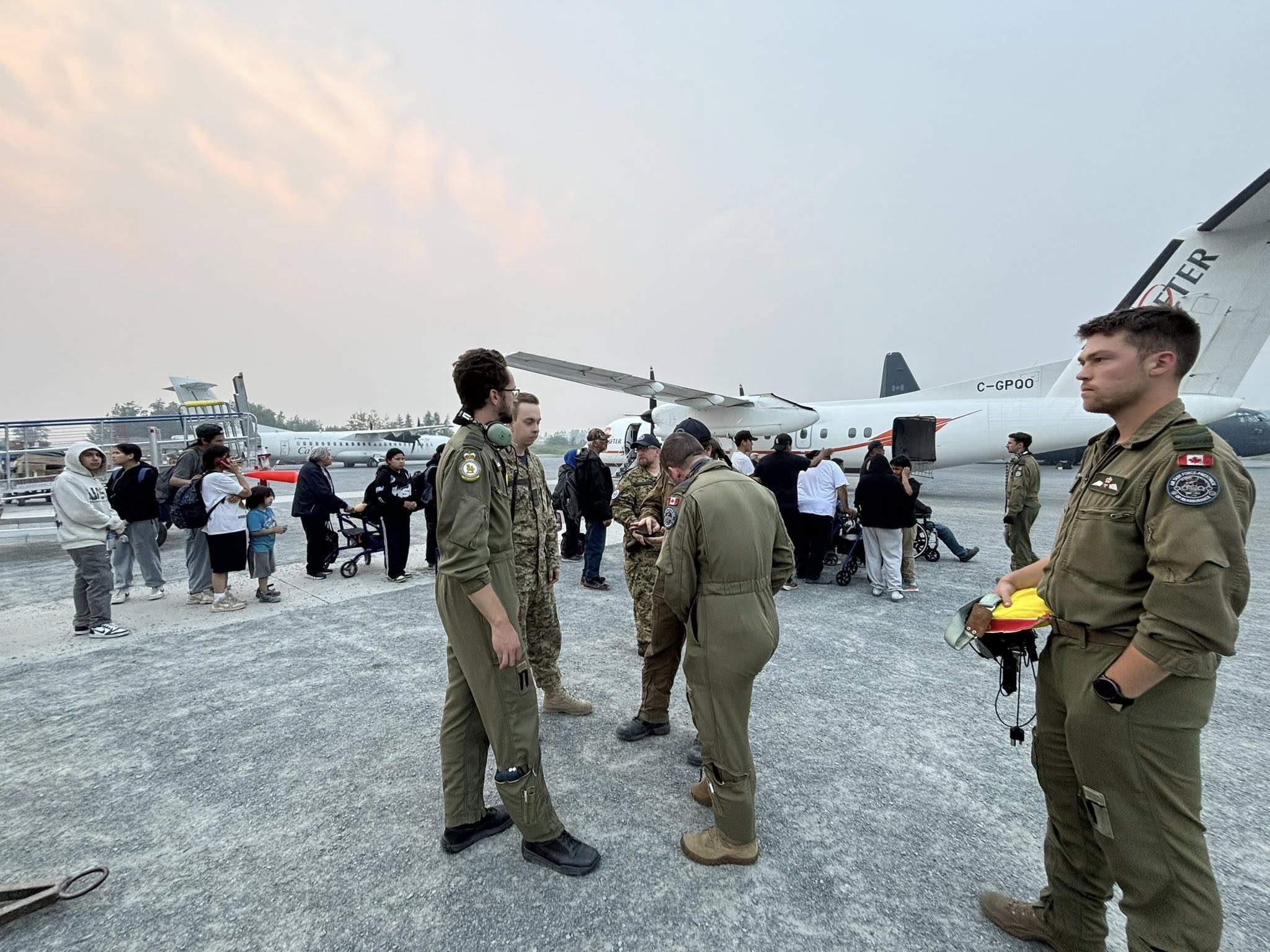 Men in brown military uniforms stand on a tarmac while a group of residents line up in the background to walk onto a large twin prop airplane